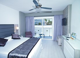 Une chambre d’hôtel moderne équipée d’un lit, d’un bureau, d’une chaise, d’un ventilateur de plafond et d’un balcon offrant une vue sur les palmiers.