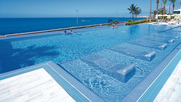 Una gran piscina infinita con agua azul cristalina, un solárium con baldosas y vistas al océano.