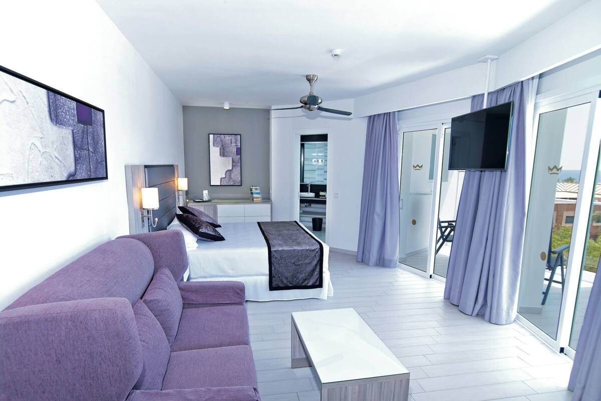 A modern hotel room with a bed, a sofa, a TV, and a balcony with curtains.