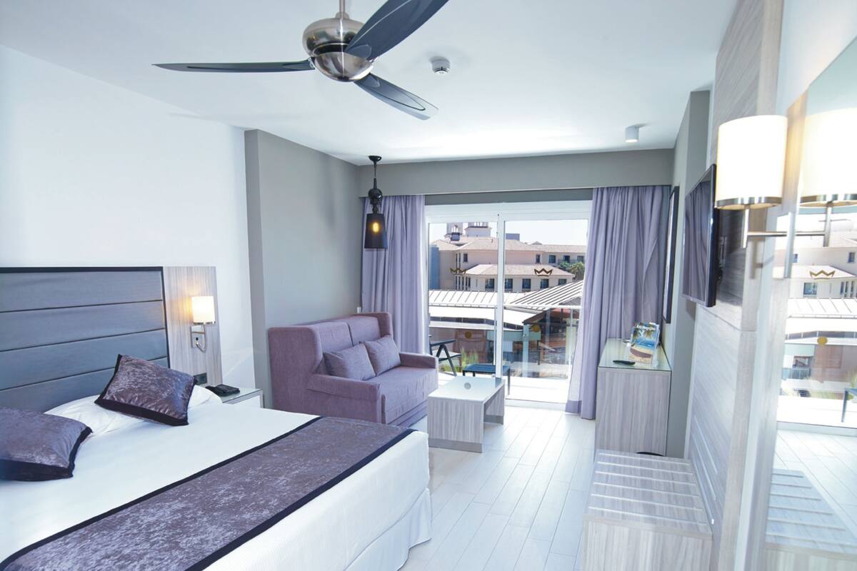 A hotel room with a bed, a sofa, a ceiling fan, a nightstand, a lamp, and a view of the outside.