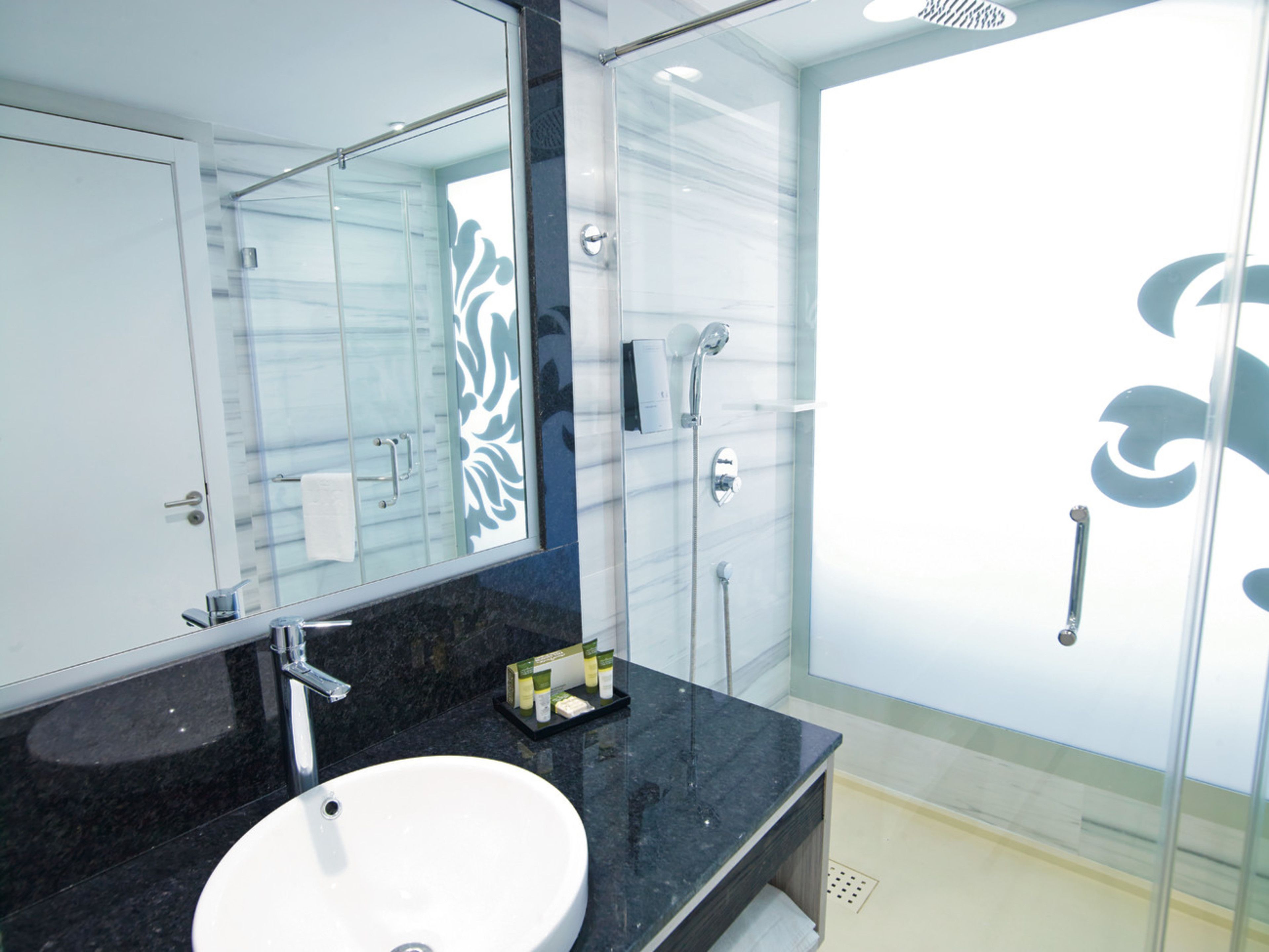 Un baño moderno con un espejo grande, un lavamanos blanco, cabina de ducha con mampara de vidrio y encimera de mármol.
