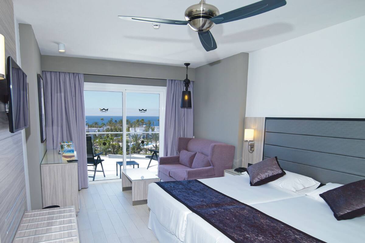 A hotel room with a bed, a desk, a chair, a balcony with a view of the sea, and a ceiling fan.