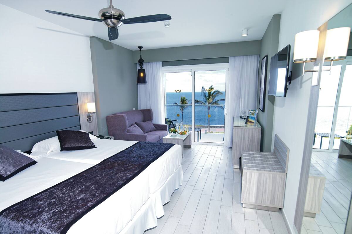 A hotel room with a bed, a chair, a table with a vase, and a balcony with a view of the sea.