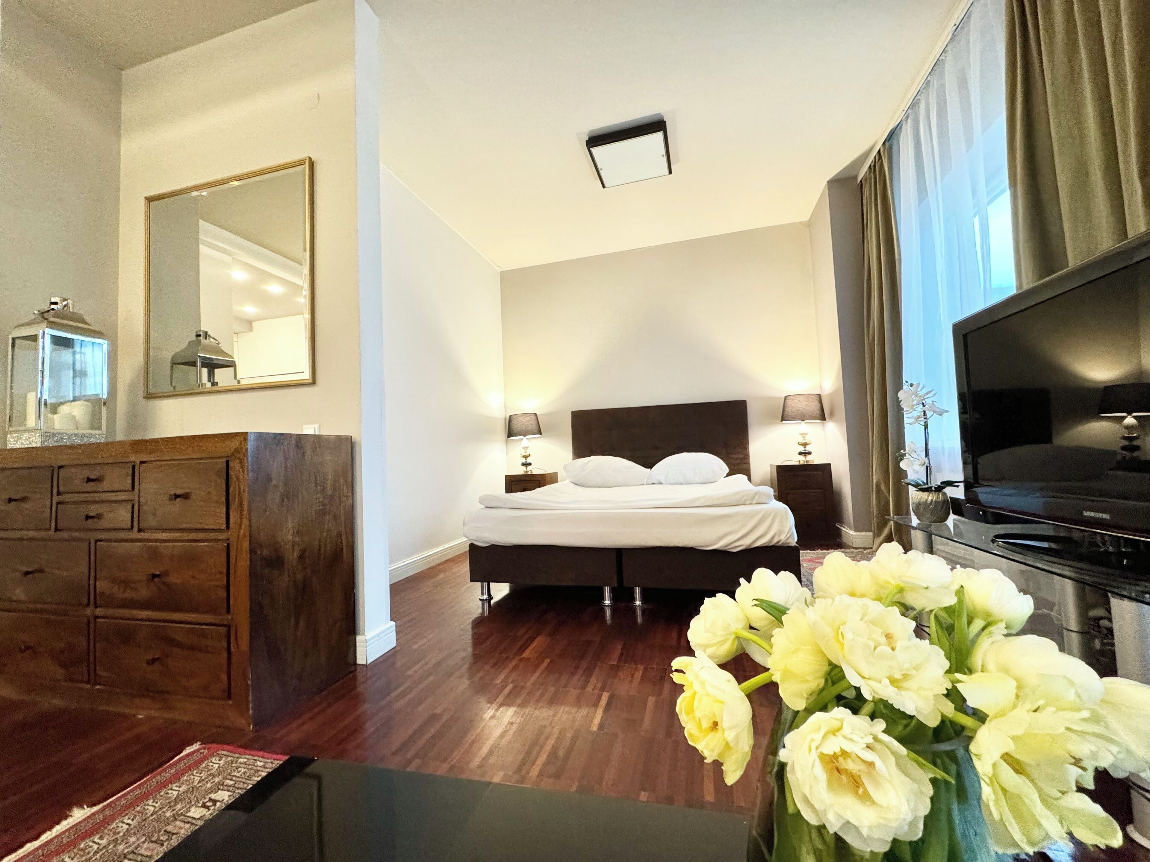 Een hotelkamer met een bed, nachtlampjes, een televisie, een dressoir, een spiegel en een bloemenvaas.