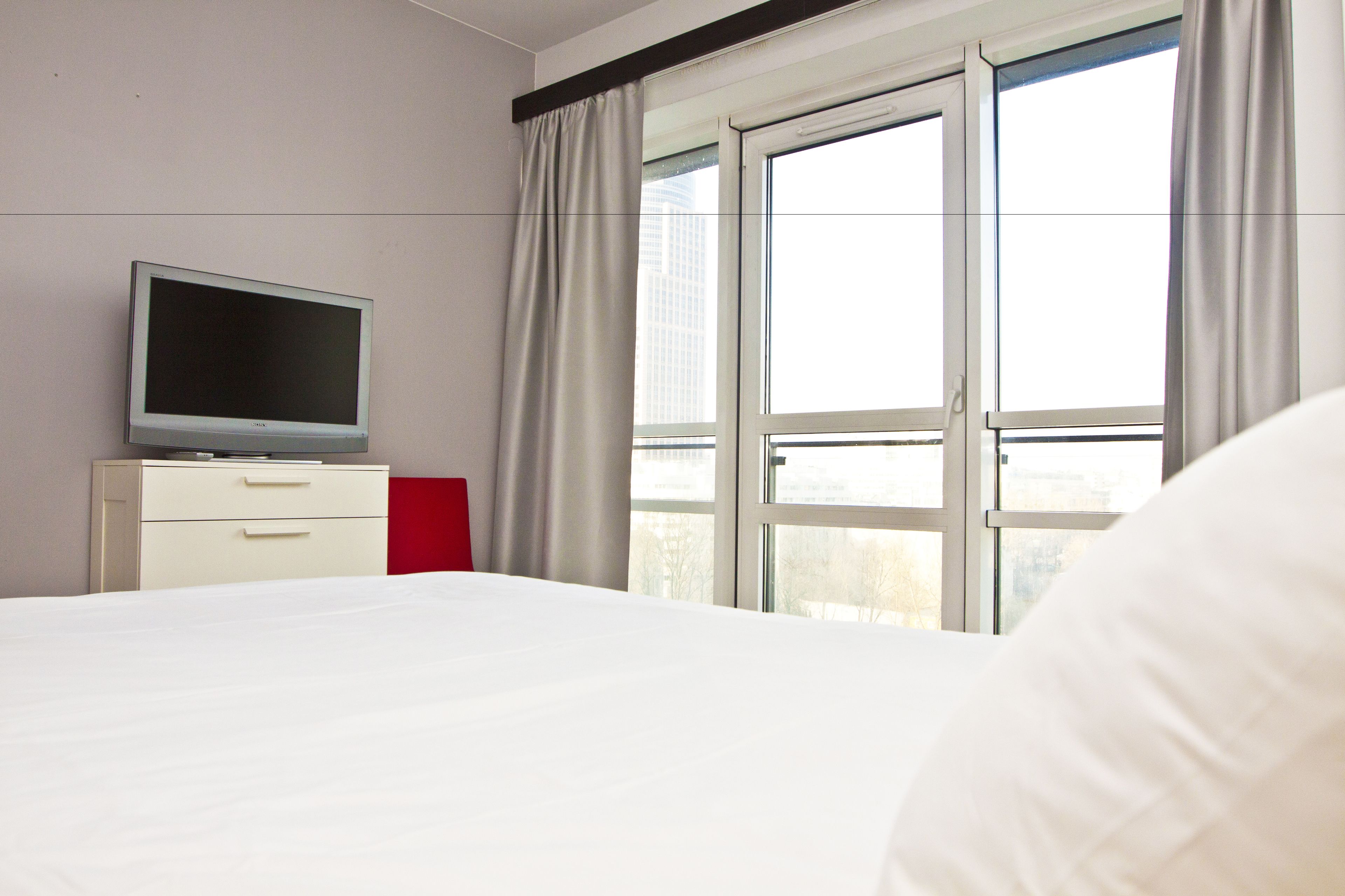 Habitación de hotel con cama, televisor en una cómoda, ventana con cortinas y vista a edificios.