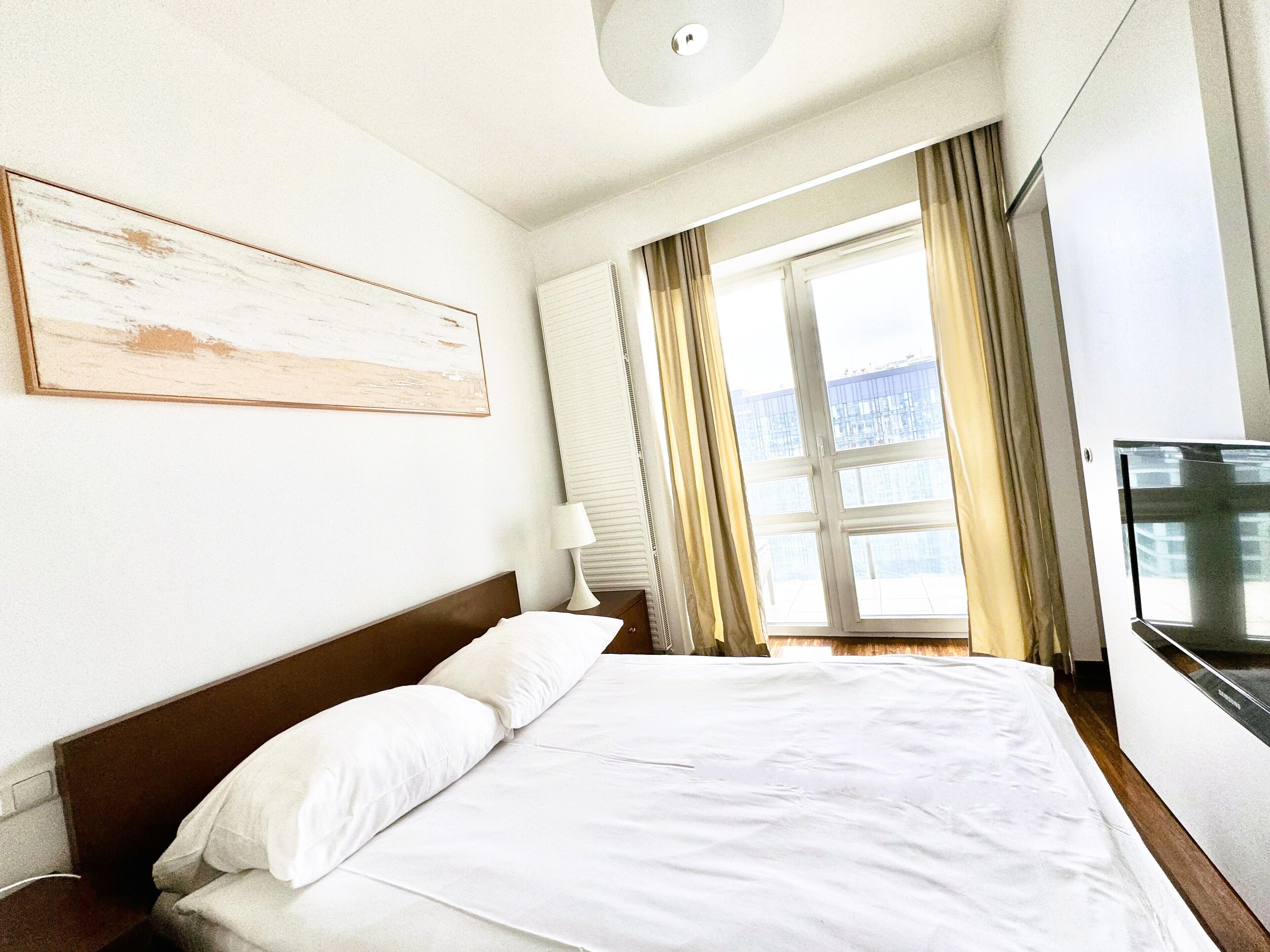 Une chambre d’hôtel avec un grand lit, une table de chevet, une lampe, une télévision et une fenêtre avec des rideaux.