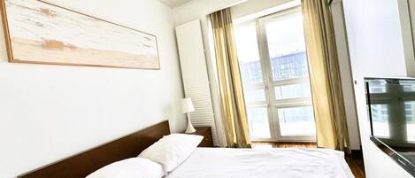 Une chambre d’hôtel avec un grand lit, une table de chevet, une lampe, une télévision et une fenêtre avec des rideaux.