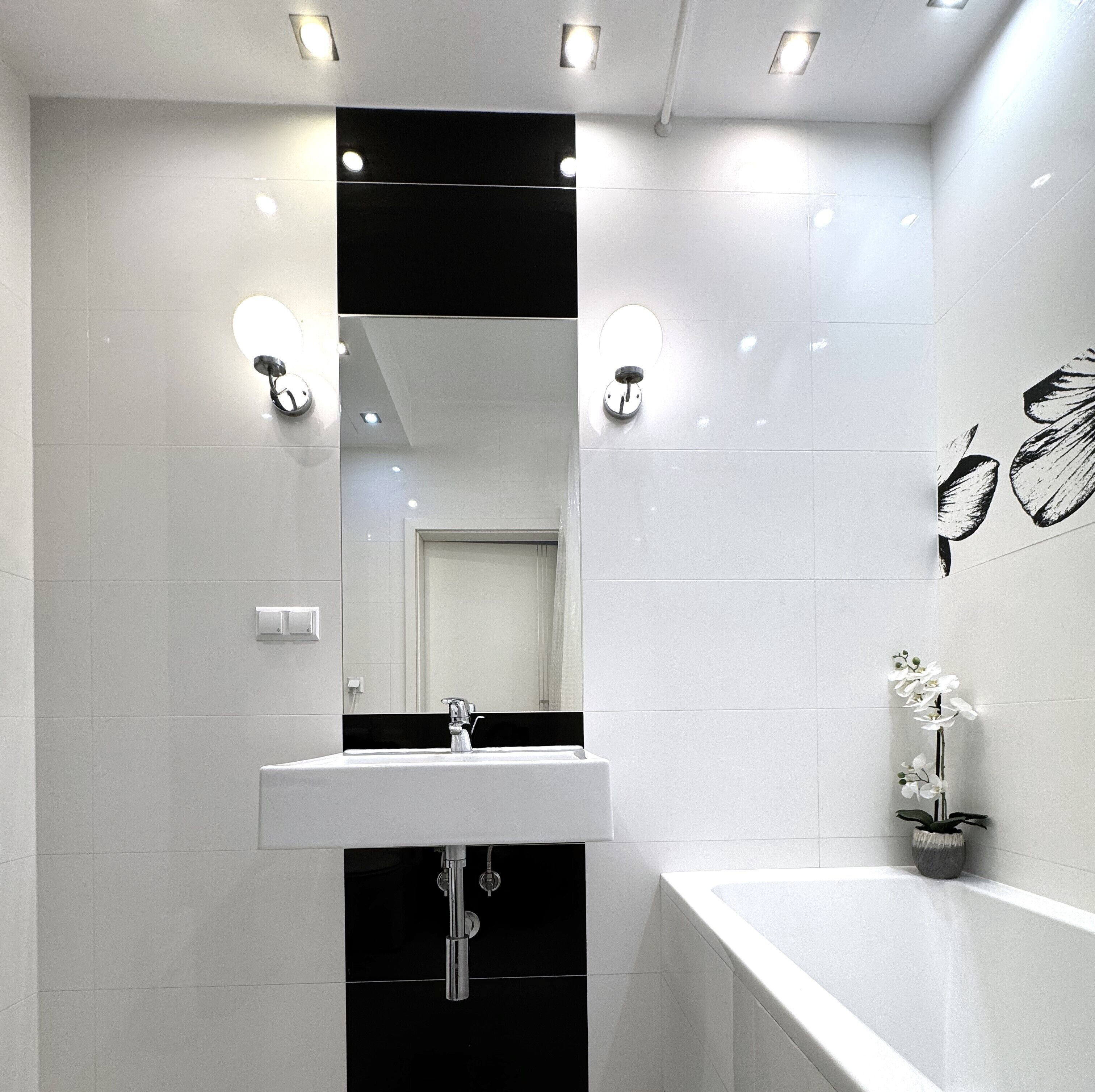 A modern bathroom with a white sink, a large mirror, and a bathtub. There are wall-mounted lights and a potted orchid on the right side.