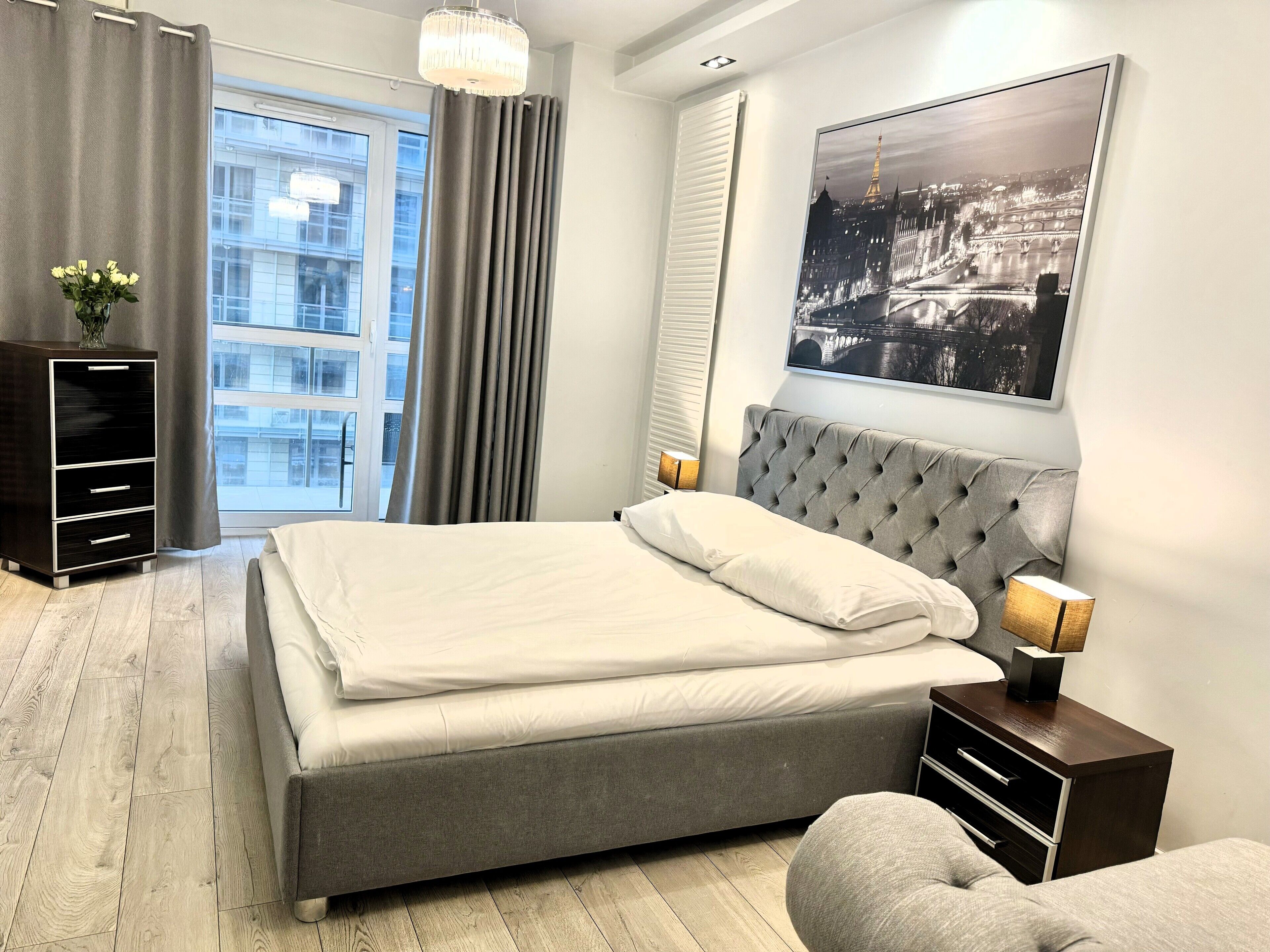A modern bedroom with a large bed, a bedside table with a lamp, a dresser, and a window with curtains.