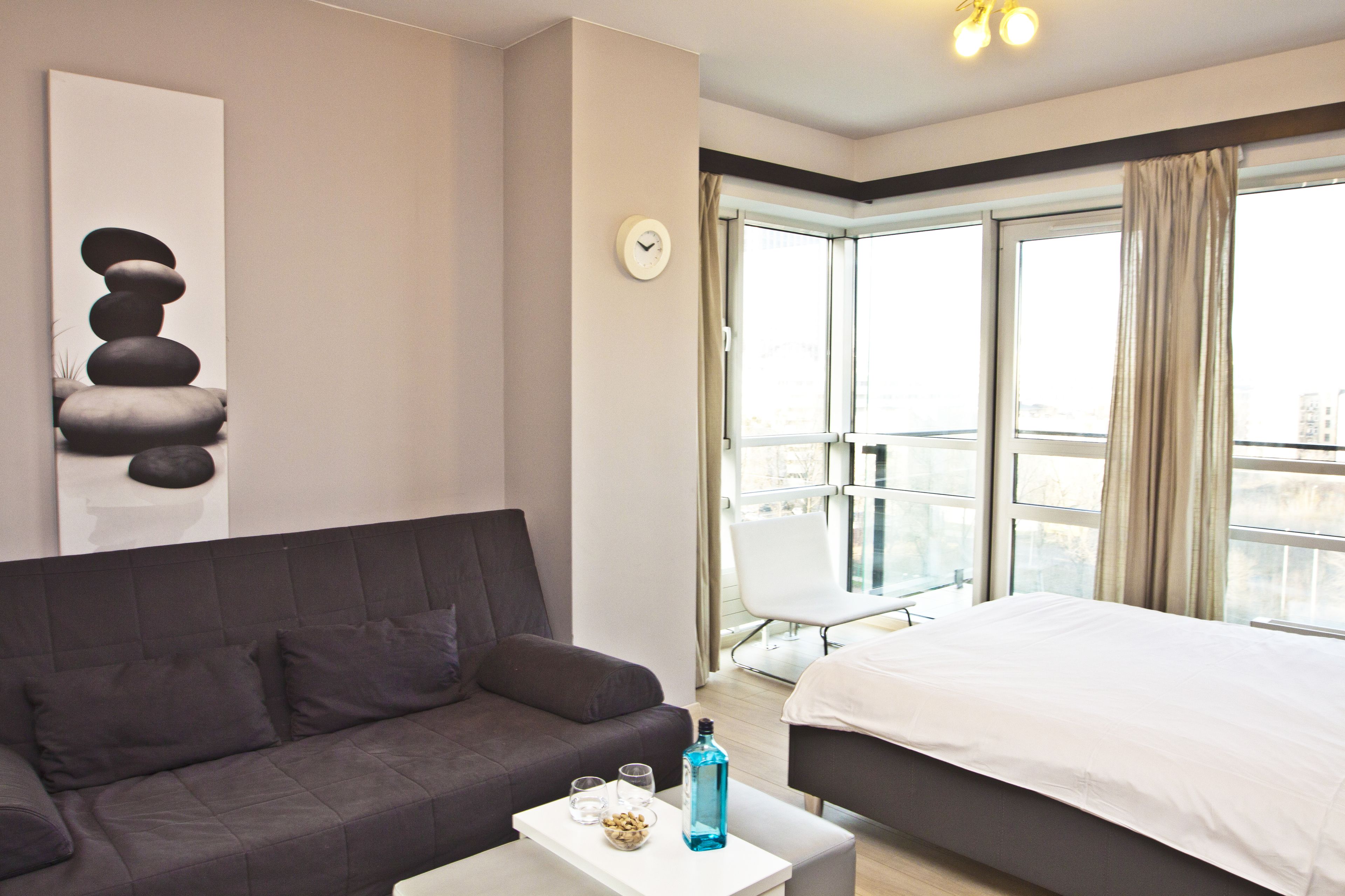 A modern hotel room with a grey sofa, a white bed, a small table with a bottle and glasses, and a balcony with a chair.