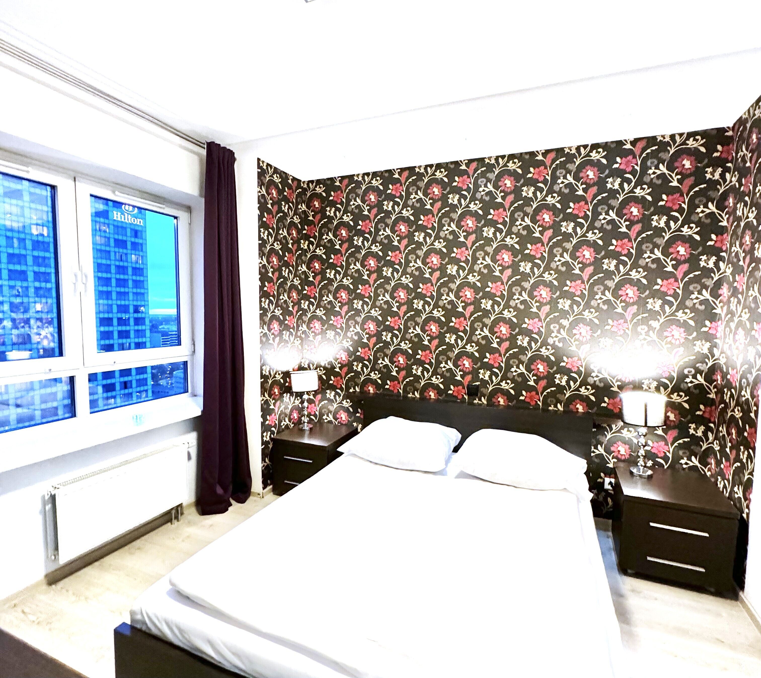 A hotel room with a large window, a floral wallpaper, a bed with white linens, and two bedside tables with lamps.