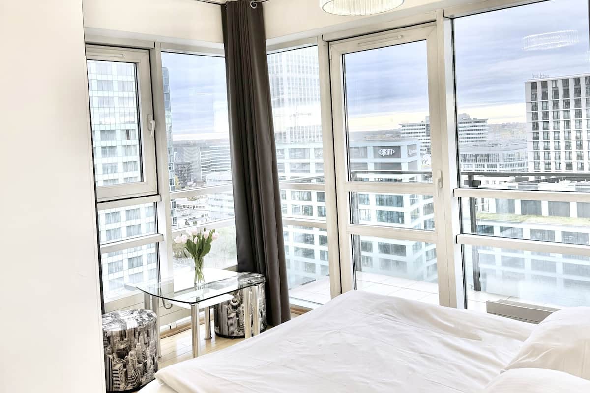Une chambre moderne avec un grand lit, une table en verre ornée d’un vase, et une vue sur la ville grâce à de grandes fenêtres.