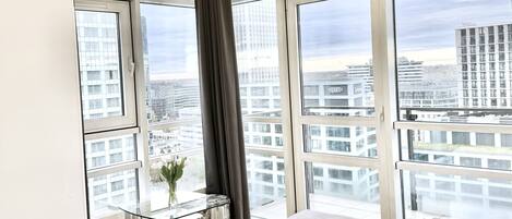 Een moderne slaapkamer met een groot bed, een glazen tafel met een vaas, en uitzicht op de stad door grote ramen.