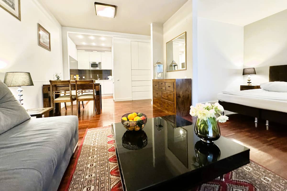 Un salon moderne avec un canapé, une table basse sur laquelle se trouve un saladier rempli de fruits, un coin repas avec une table et des chaises en bois, et une cuisine en arrière-plan.