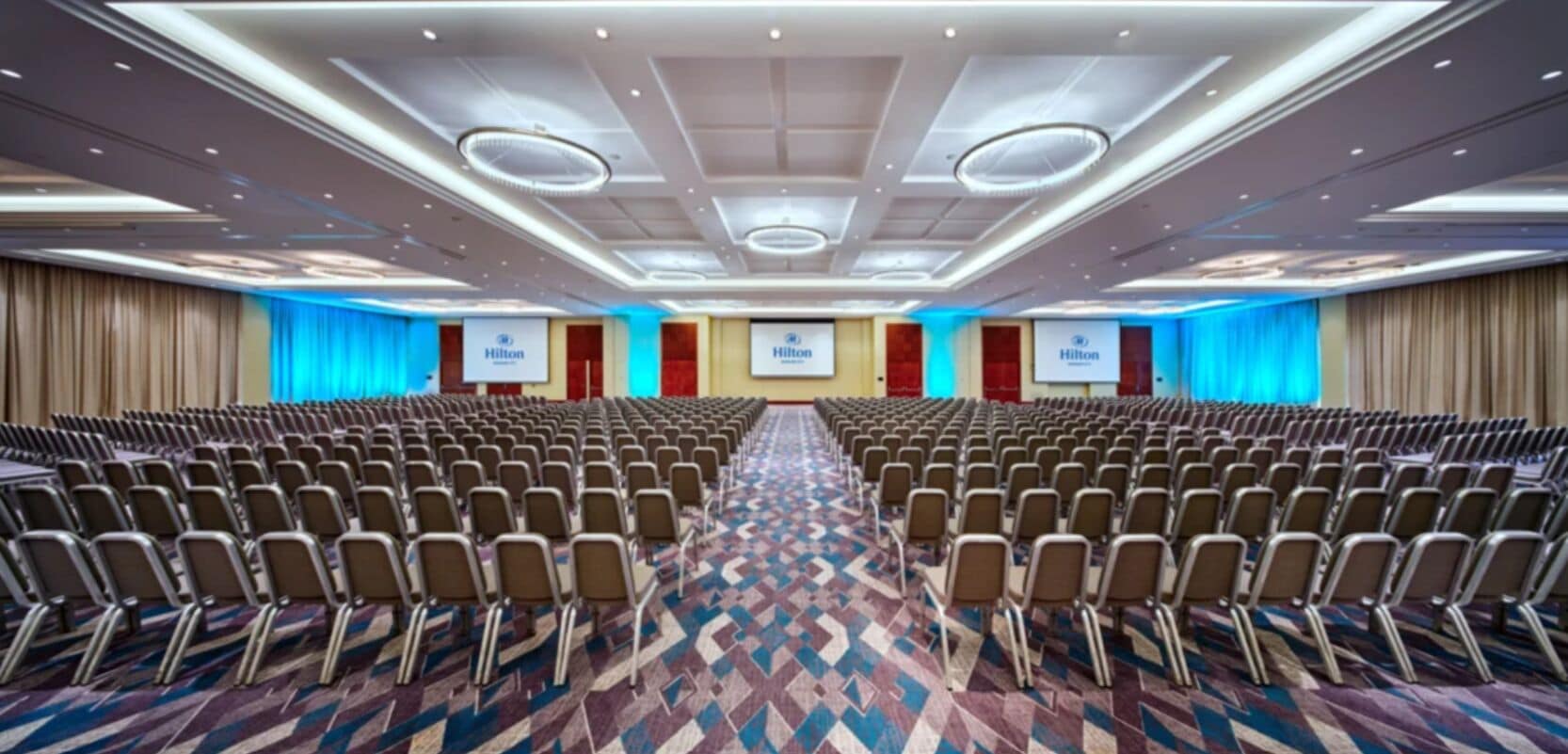 A large conference room with rows of chairs, a patterned carpet, and a ceiling with recessed lighting.
