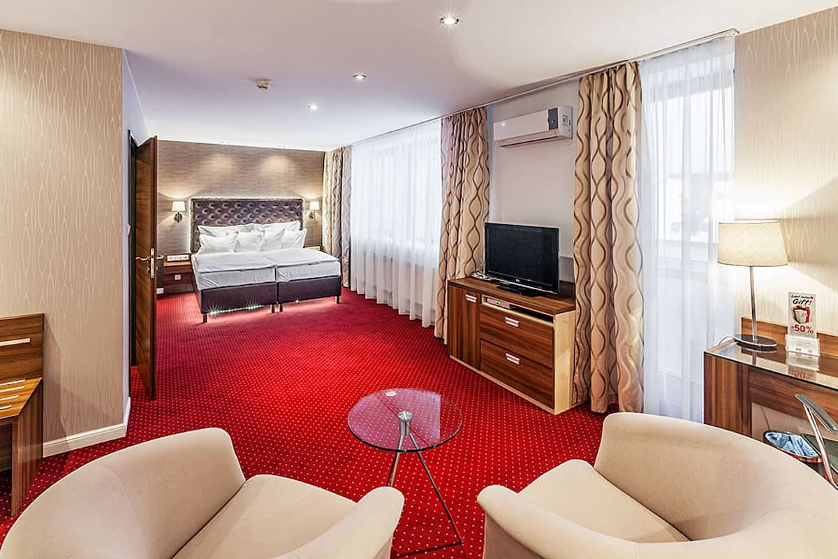 A hotel room with a red carpet, a bed with a headboard, a TV, a desk with a lamp, and two armchairs.