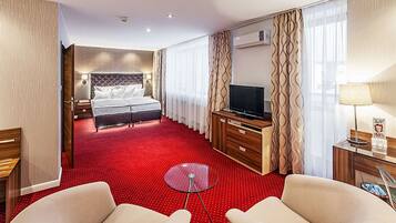 A hotel room with a red carpet, a bed with a headboard, a TV, a desk with a lamp, and two armchairs.
