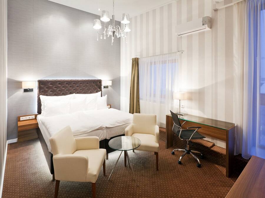 A hotel room with a large bed, two armchairs, a glass table, a desk with a chair, a chandelier, and a window with curtains.