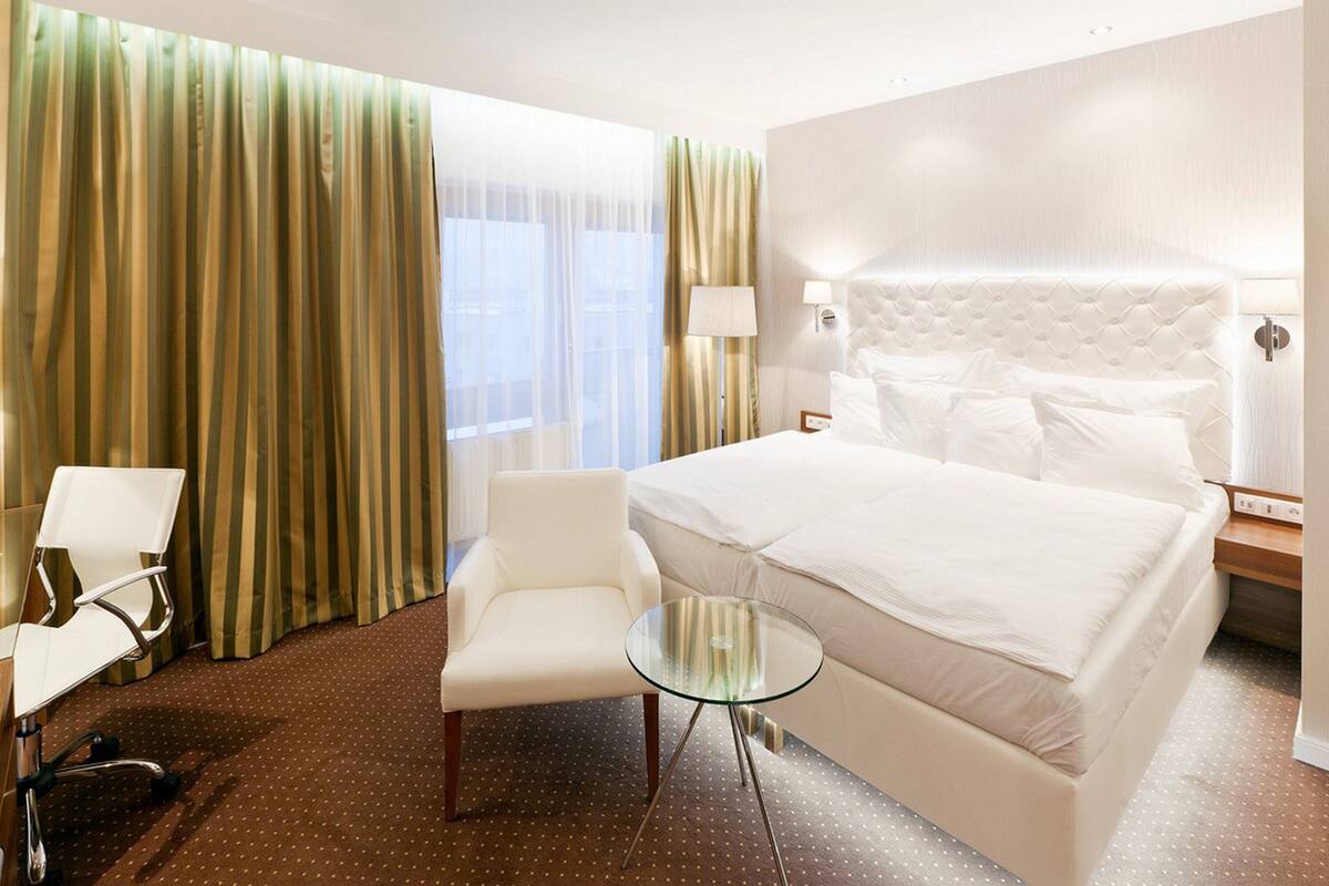 A hotel room with a large bed, a chair, a glass table, and curtains.