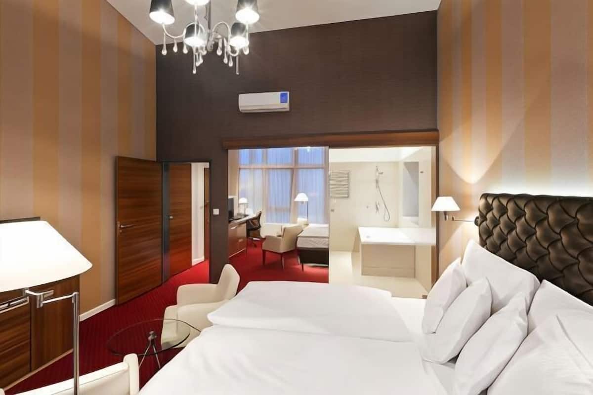 A hotel room with a large bed, a desk, a chair, a small table, and a bathroom visible through an open door.