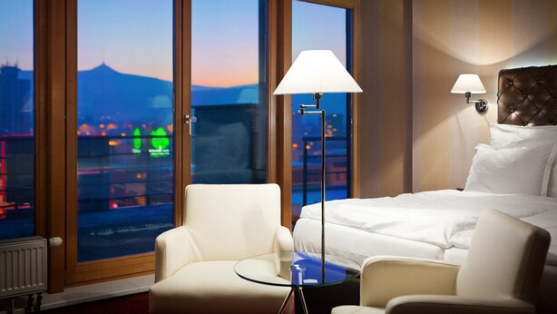 A hotel room with a large window, a bed, two chairs, a small table, and two wall-mounted lamps.