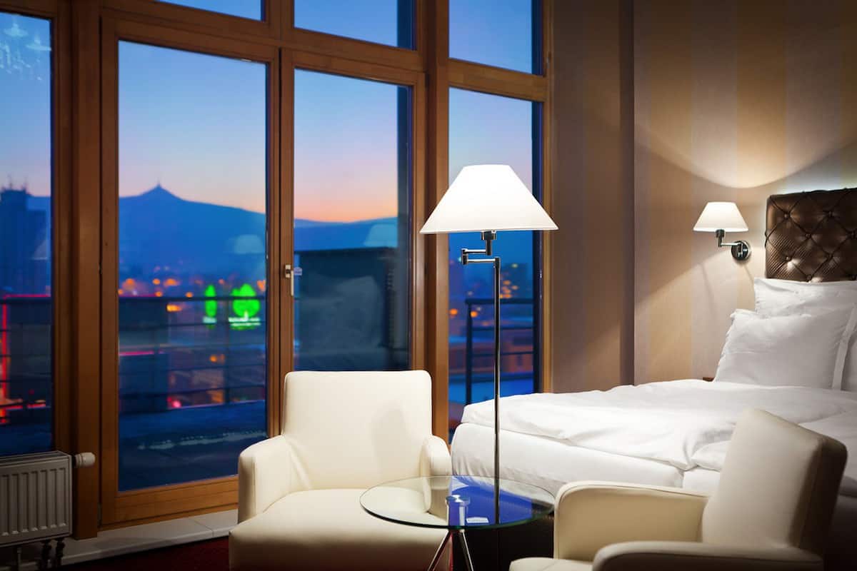 A hotel room with a large window, a bed, two chairs, a small table, and two wall-mounted lamps.
