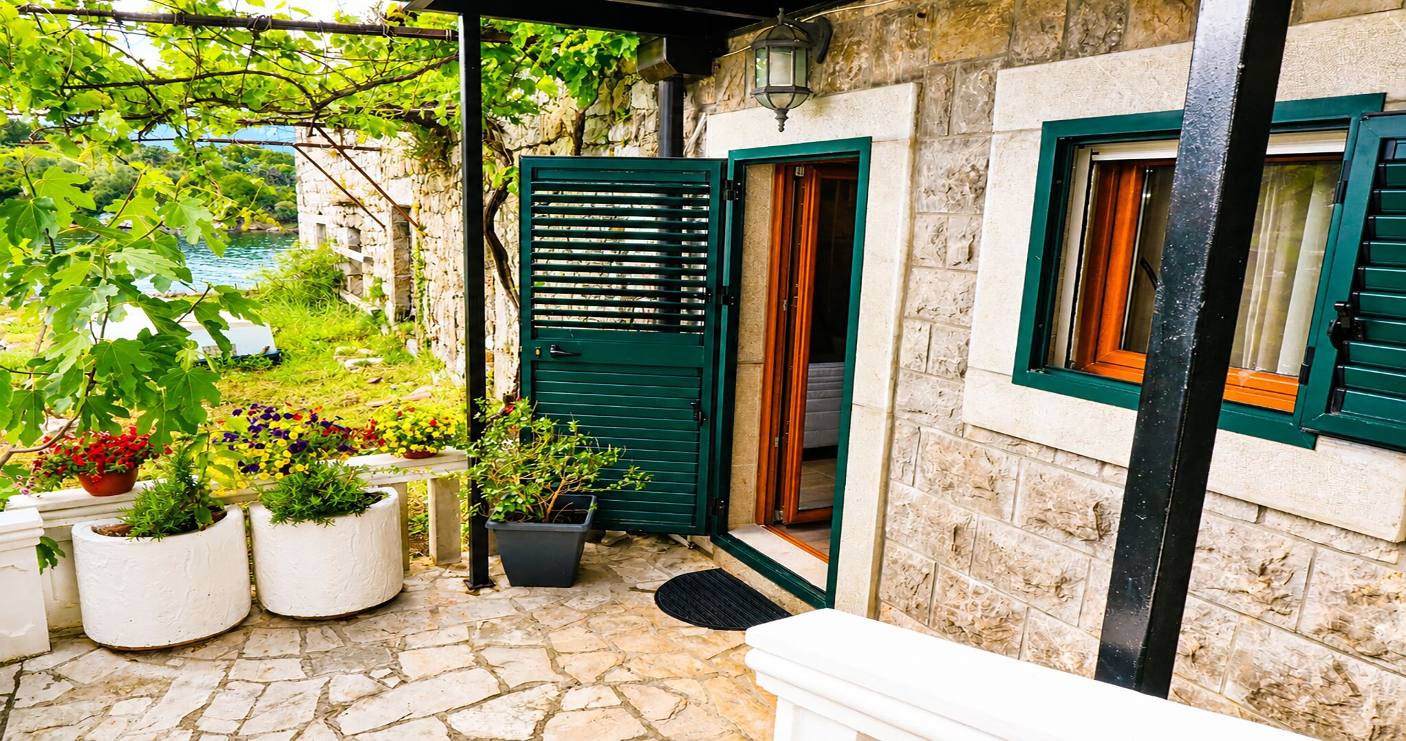 A stone building with a green door and windows, a stone patio with potted plants, and a view of greenery and a body of water.
