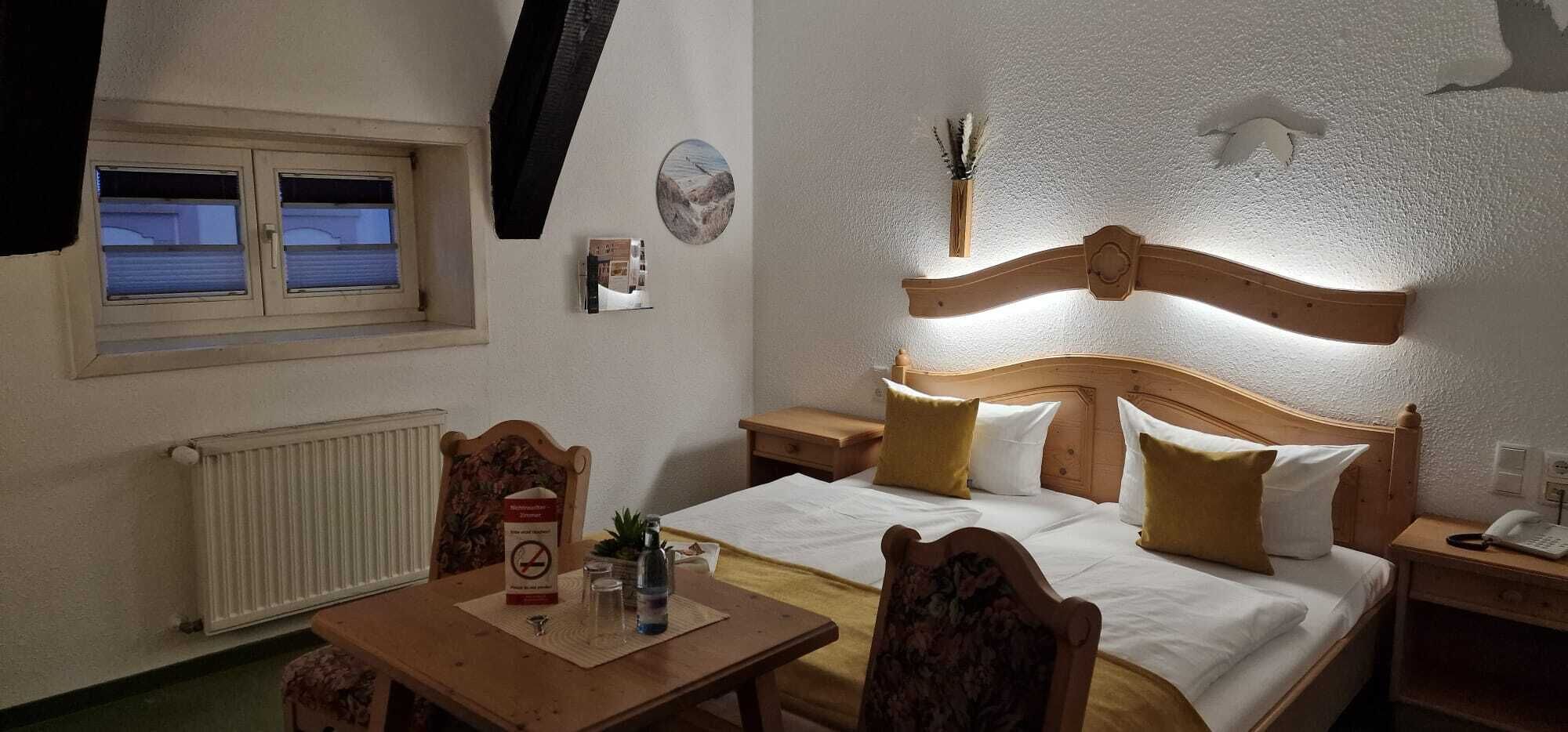 Una habitación de hotel con cama, mesitas de noche, una mesa con sillas, una ventana y un radiador.