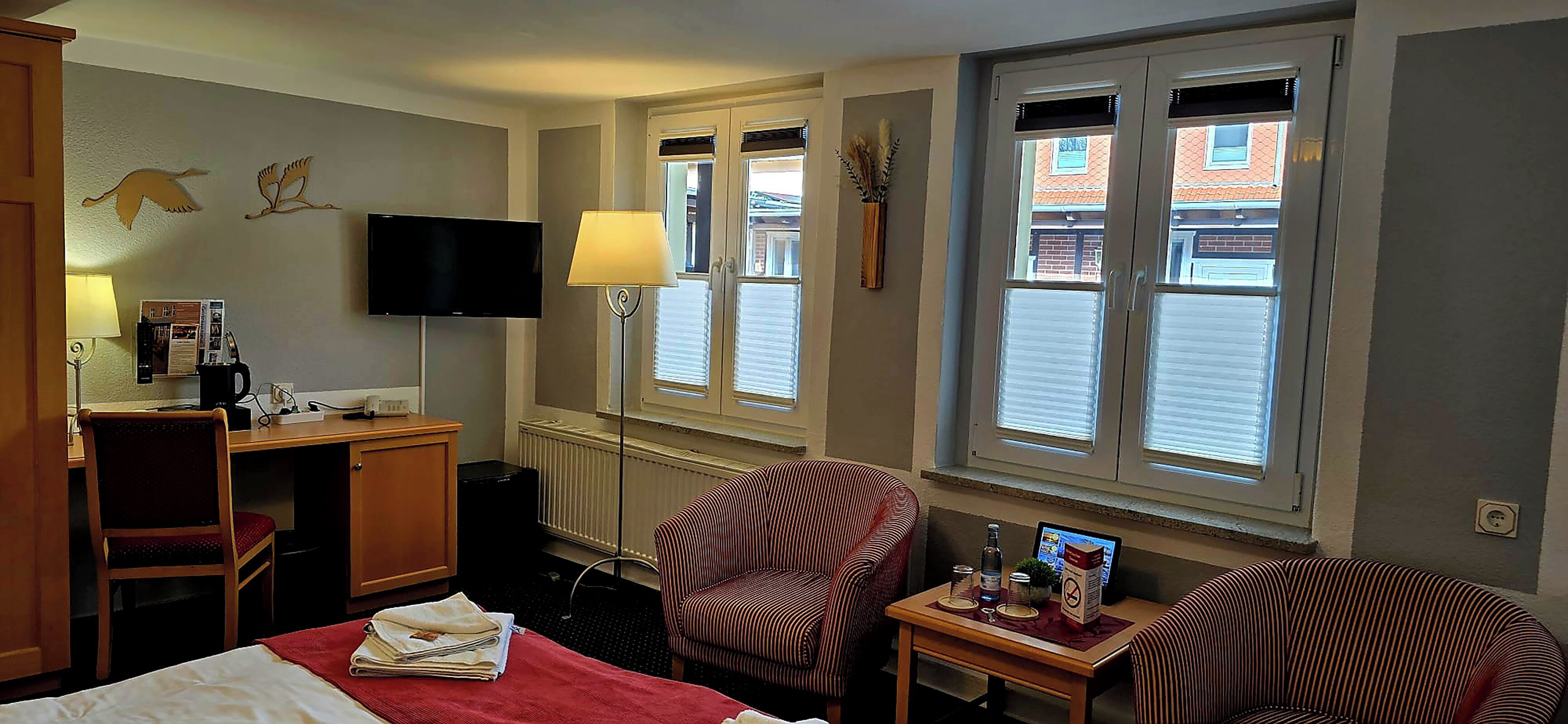 A hotel room with a bed, a desk, a chair, a television, a lamp, a window, and a small table with a bottle and a book.
