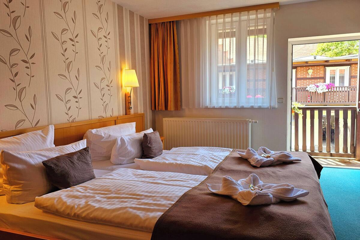 A double bed with a wooden headboard, a bedside table with a lamp, a window with curtains, and a view of a balcony with flowers.