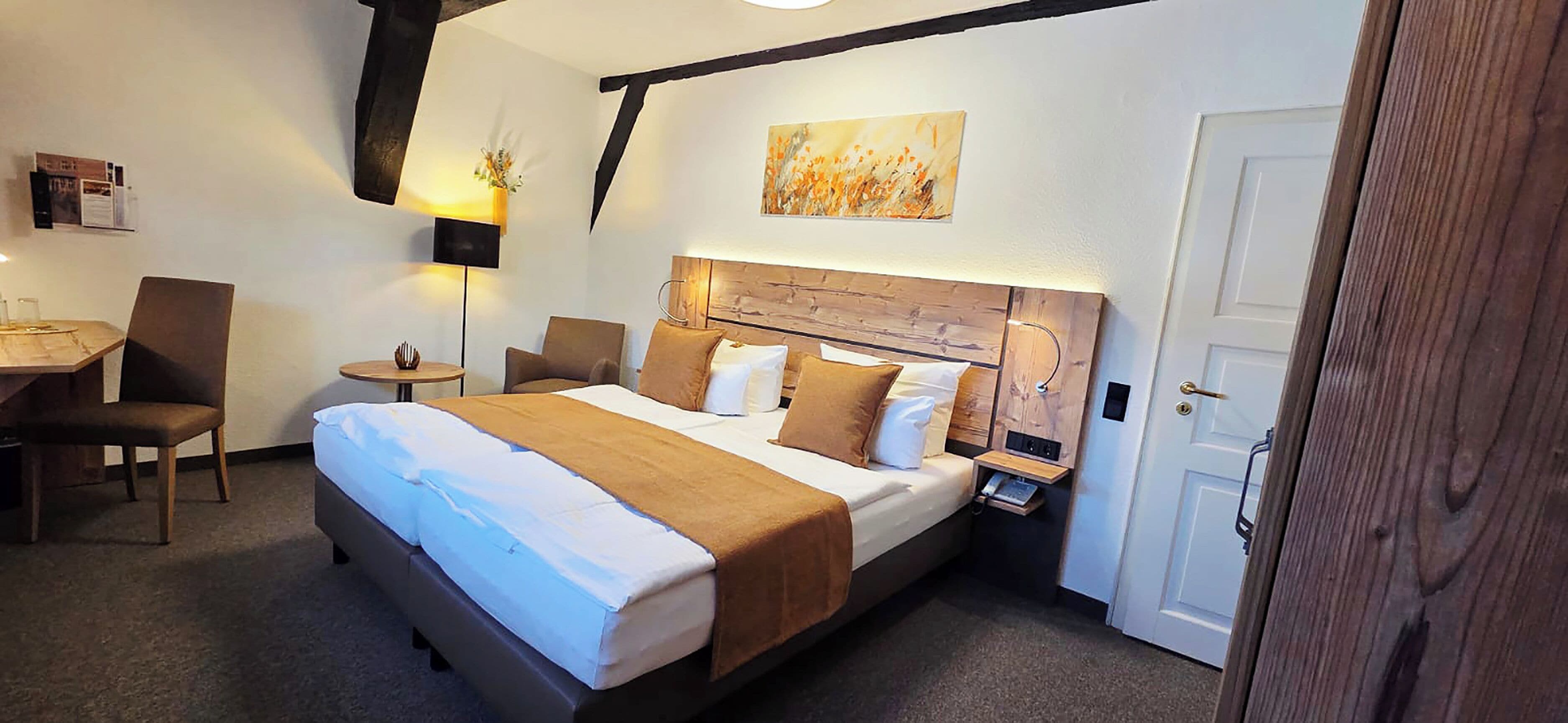 A hotel room with a large bed, a desk with a chair, a bedside table with a lamp, and a wooden headboard.