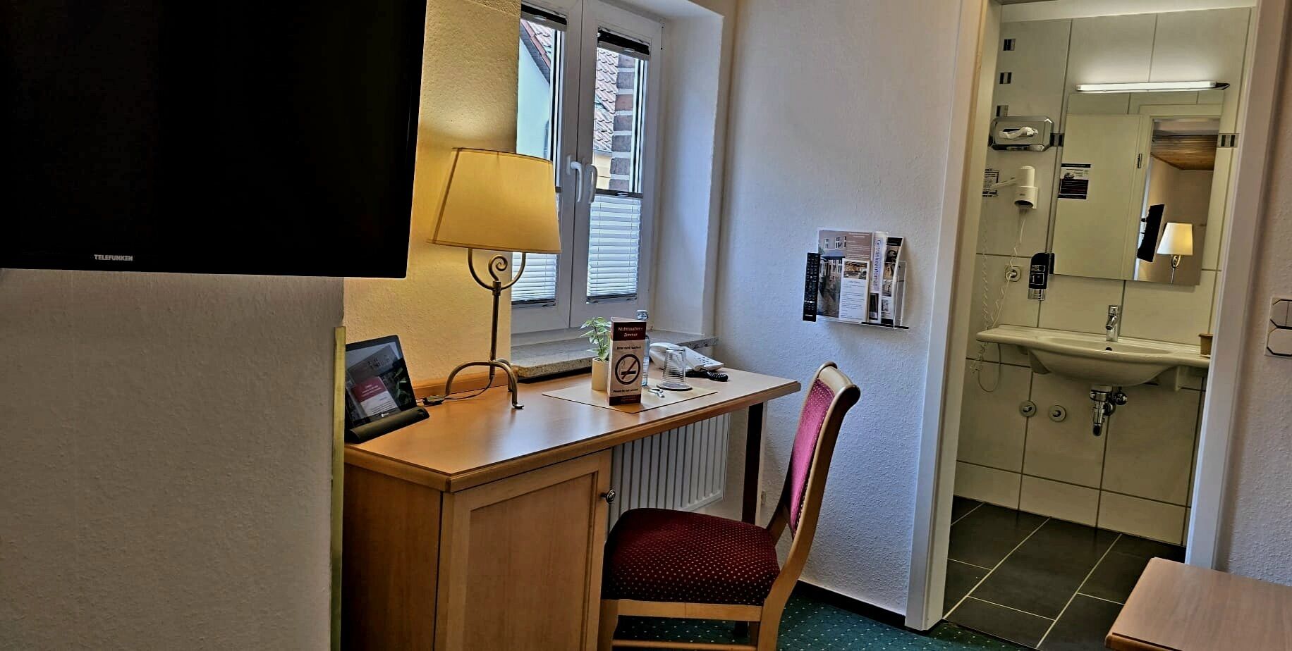 Une chambre d’hôtel équipée d’un bureau, d’une chaise, d’une télévision, d’une fenêtre avec stores et d’une salle de bain avec un lavabo et un miroir.