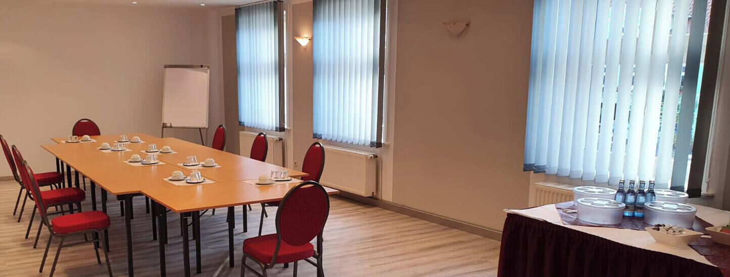 Een vergaderruimte met een lange tafel, rode stoelen, een whiteboard en een buffet met eten en drinken.