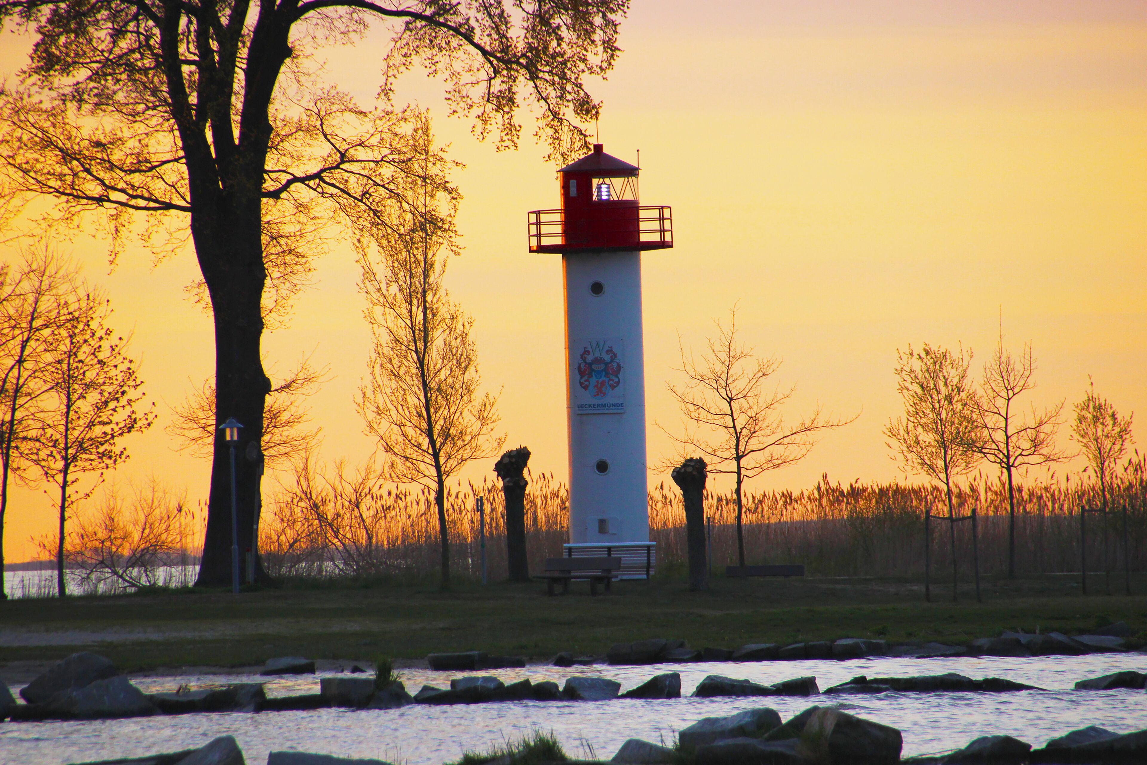 Un faro con la parte superior roja, situado junto a un río con rocas en primer plano y árboles al fondo.