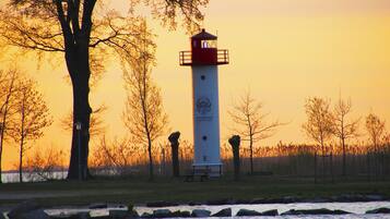 Un faro con la parte superior roja, situado junto a un río con rocas en primer plano y árboles al fondo.
