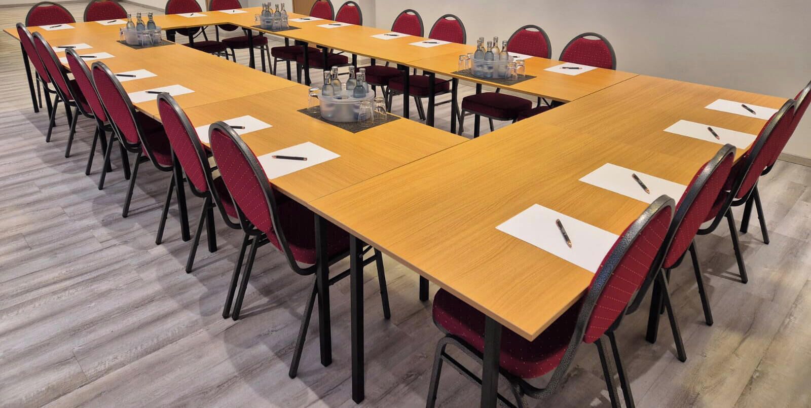 Een vergaderzaal met lange tafels, rode stoelen en waterflessen.