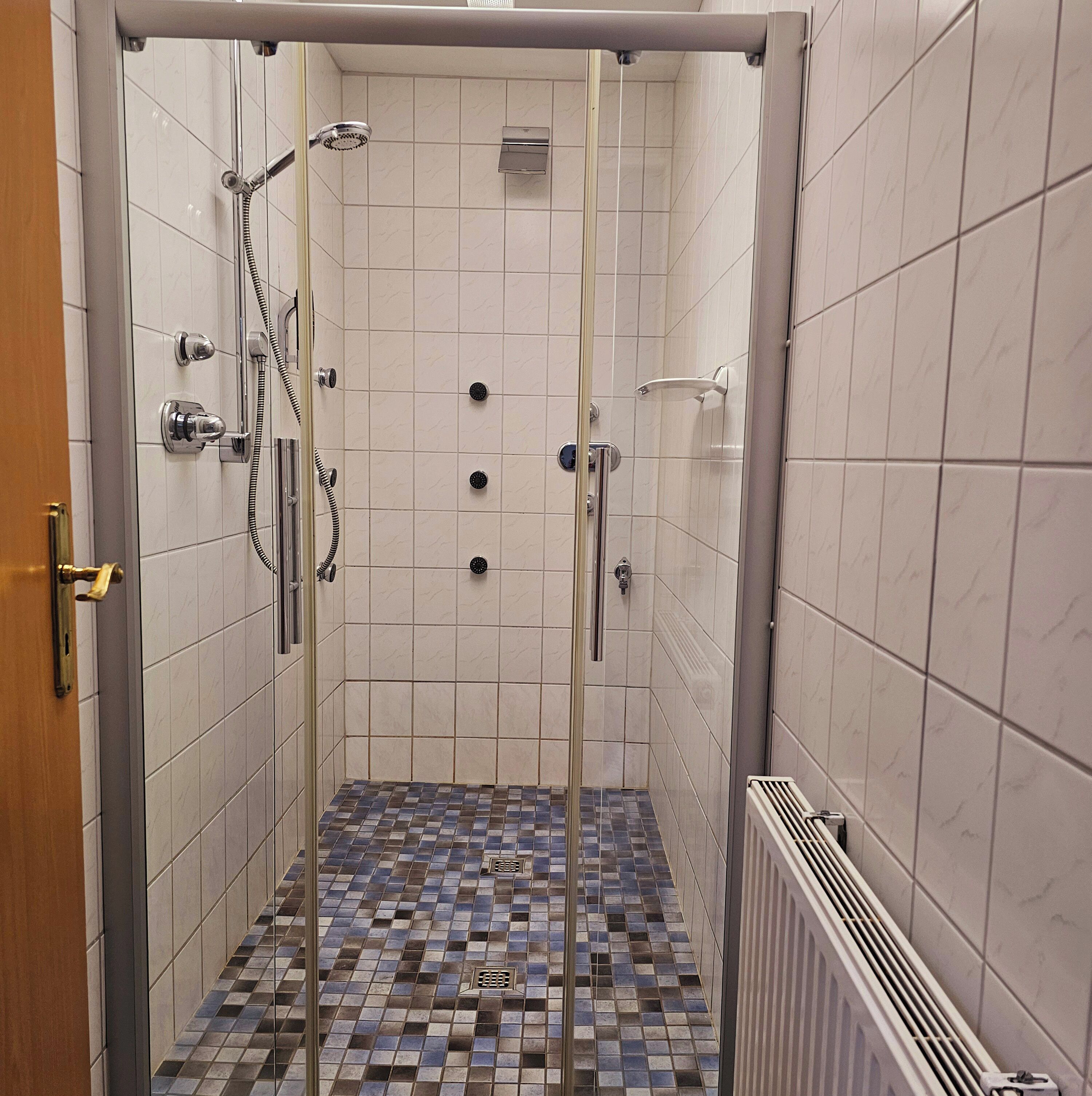 Un espace douche doté d’une porte en verre, de murs recouverts de carreaux blancs et d’un sol à motifs.