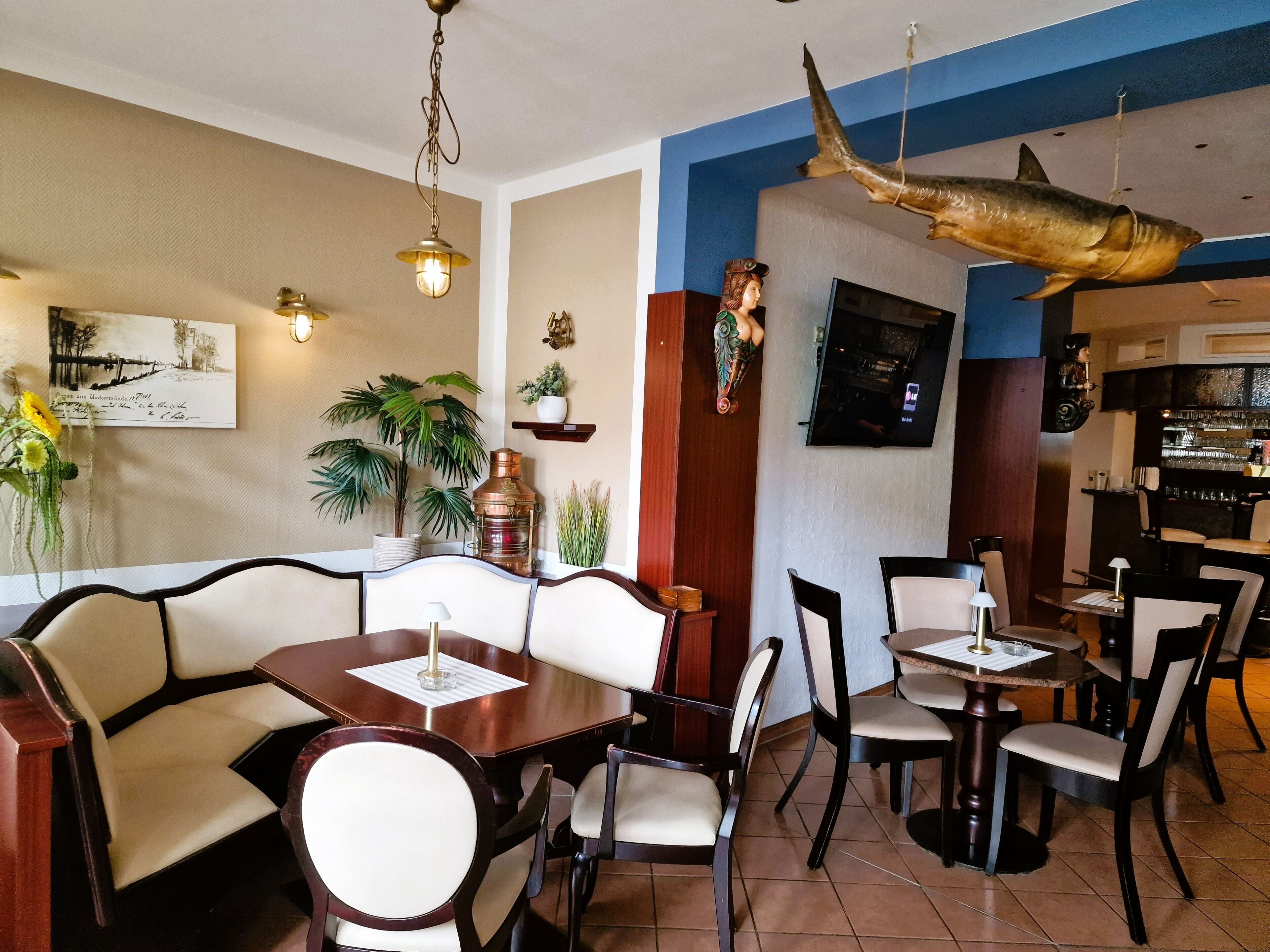A restaurant interior with wooden tables and chairs, a hanging fish decoration, and potted plants.
