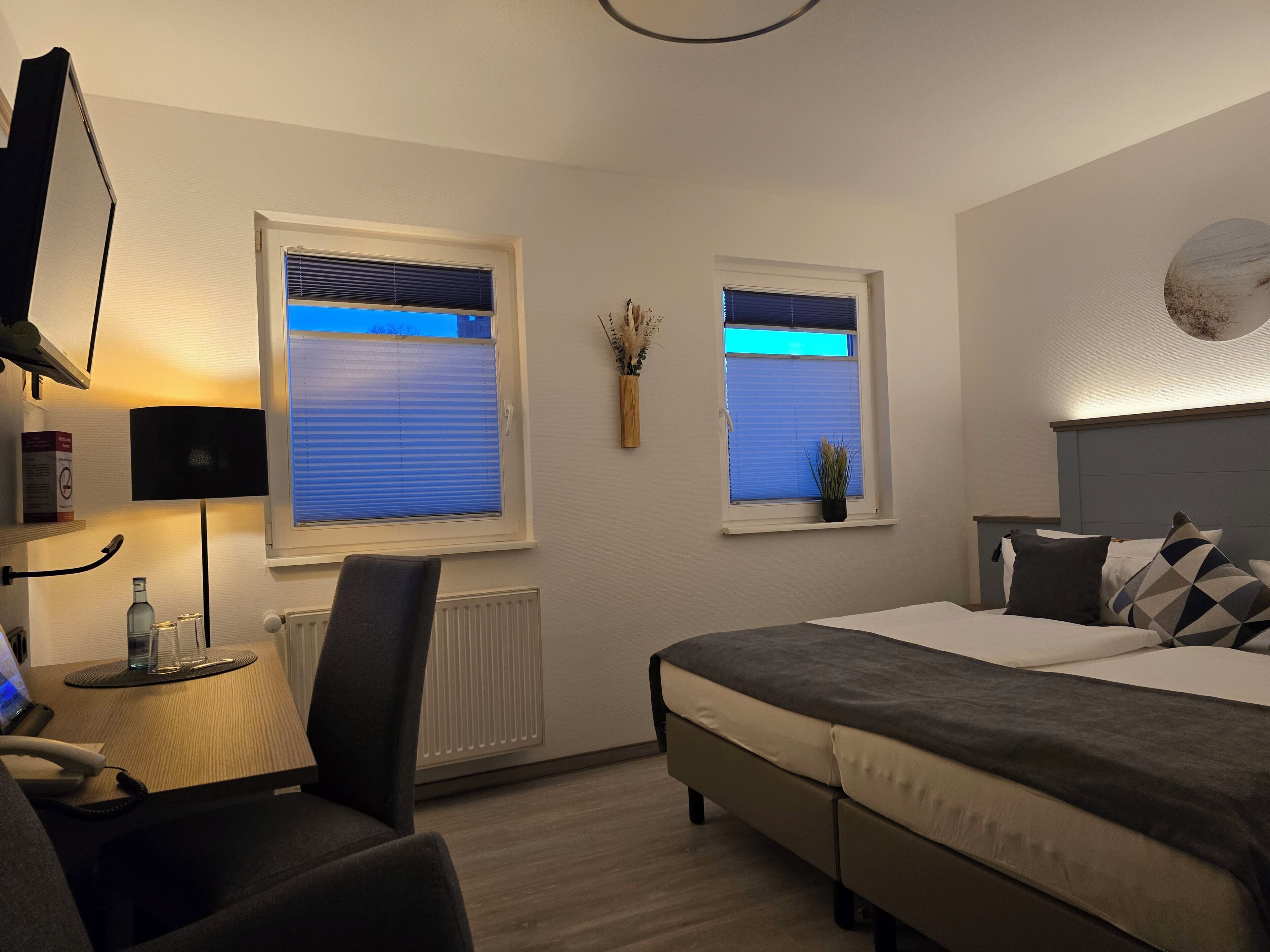 A hotel room with a bed, a desk with a chair, a radiator, two windows with blinds, a wall-mounted TV, and a small table with a lamp.