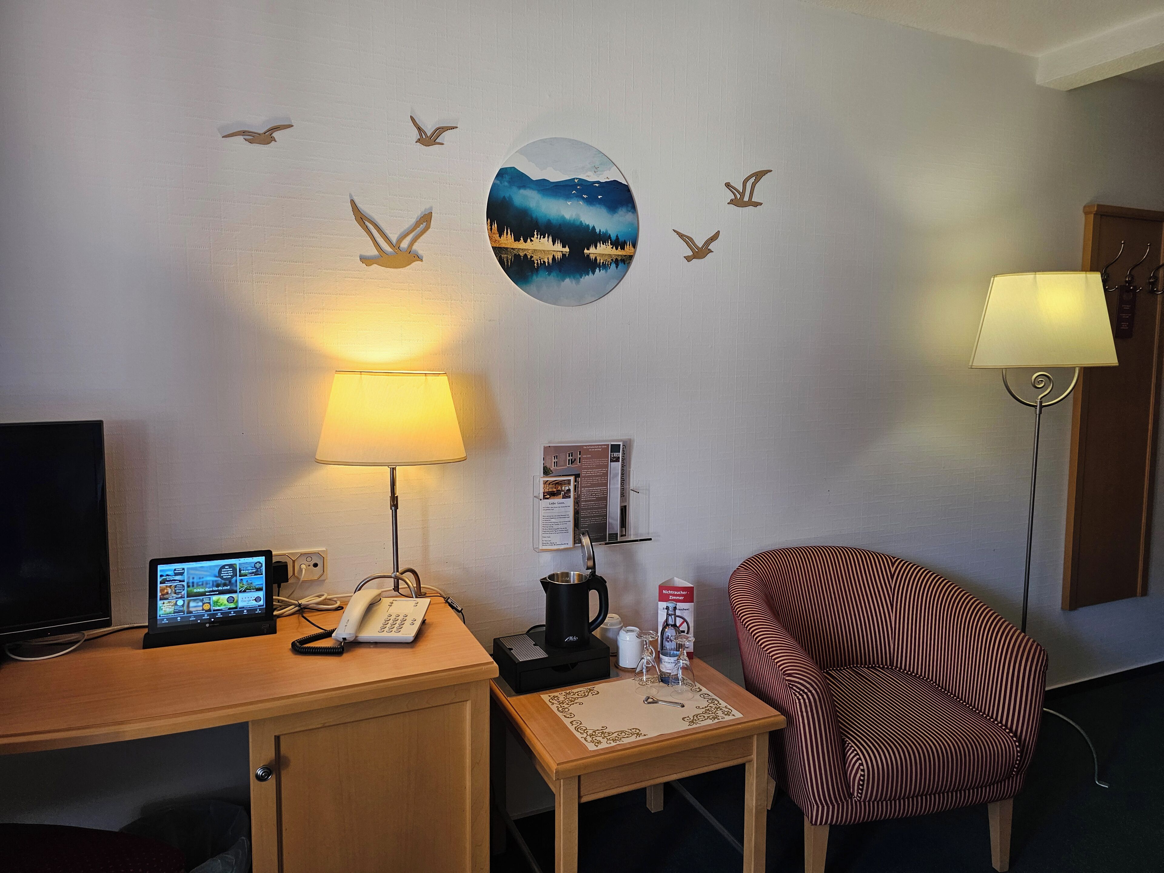 Ein Hotelzimmer mit Schreibtisch, Stuhl, Lampe, Kaffeemaschine und Wanddekorationen, darunter Vögel und ein Landschaftsbild.
