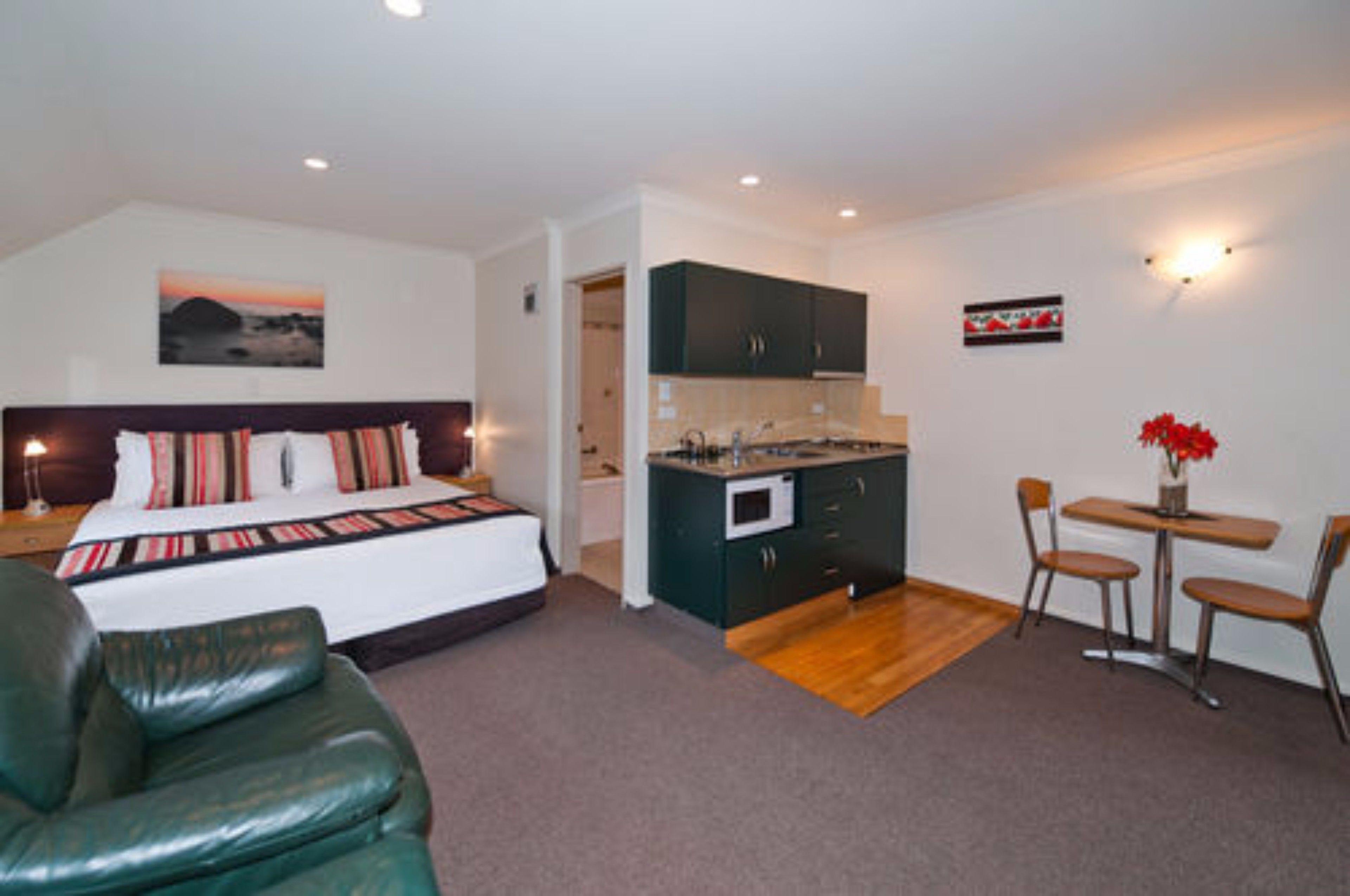 A bedroom with a bed, a green leather chair, a small kitchenette, and a dining area with a table and chairs.