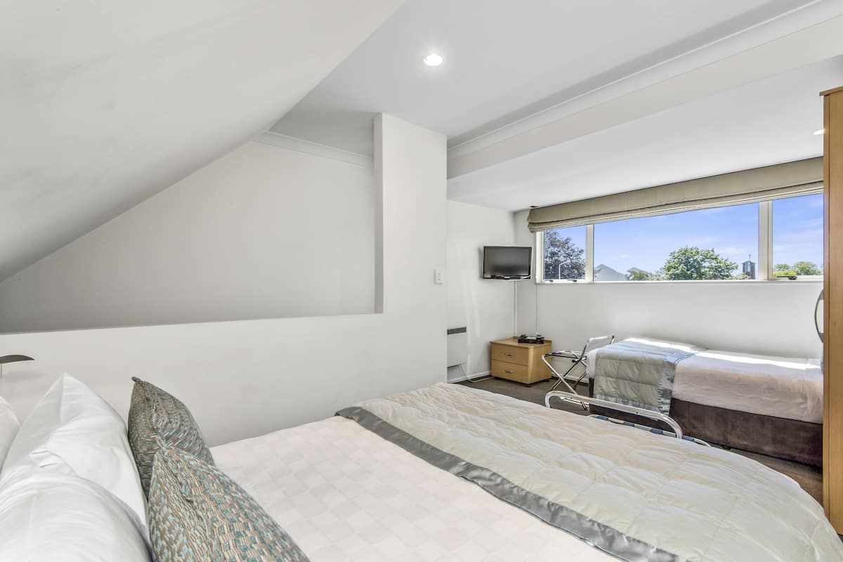 A modern bedroom with a sloped ceiling, a large bed with multiple pillows, a bedside table, a TV mounted on the wall, and a window with a view of the outside.