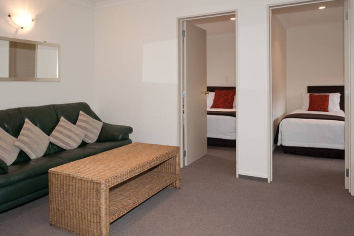 A hotel room with a green sofa, a wicker bench, and two beds with red pillows.