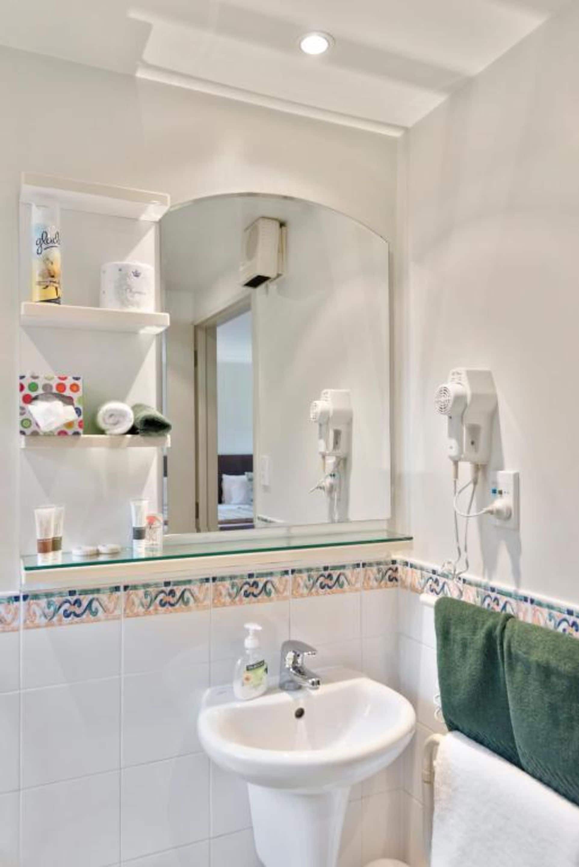 A bathroom with a glass shelf holding toiletries, a white sink, a towel rack with a green towel, and a wall-mounted hairdryer.