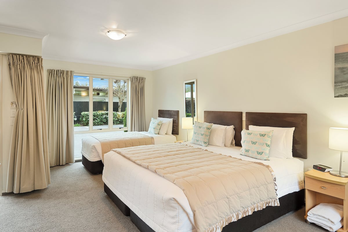 A hotel room with two beds, beige curtains, a large window, and a nightstand with a lamp.