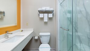A bathroom with a white sink, a toilet, a glass shower enclosure, and a towel rack with folded towels. - Cardinal Motel (Bowling Green)