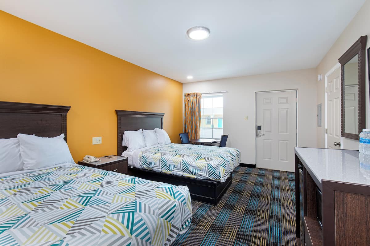 Quarto de hotel com duas camas, uma escrivaninha, uma cadeira, banheiro e um espelho grande.