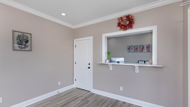 A room with a wooden floor, a white door, a framed floral artwork, a shelf with decorative items, and a wall-mounted mirror. - Cardinal Motel (Bowling Green)