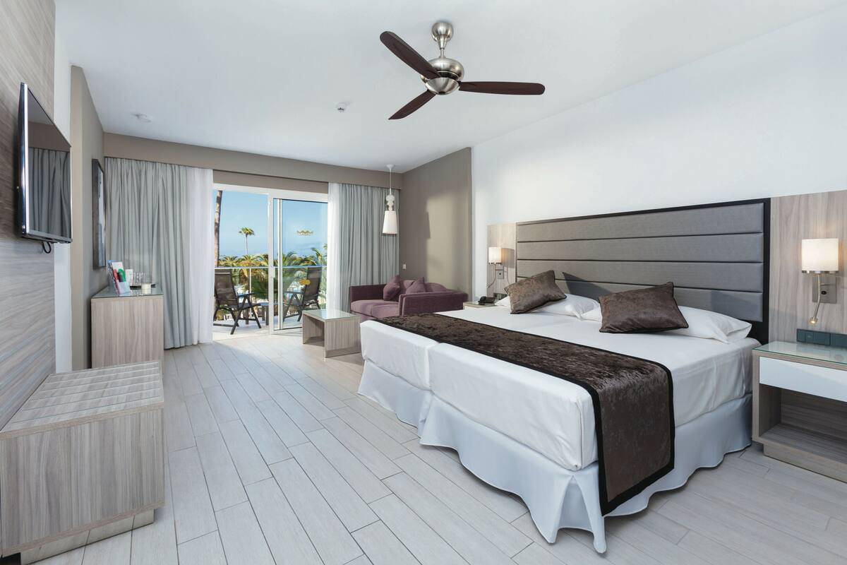 A modern hotel room with a large bed, a ceiling fan, a TV mounted on the wall, a balcony with a view, and a small desk area.