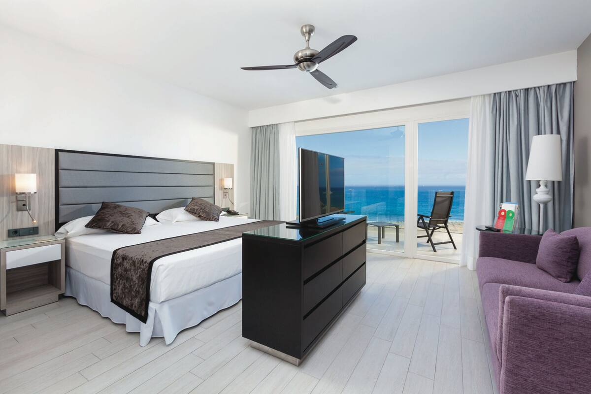 A modern hotel room with a bed, a desk, a chair, and a view of the ocean.